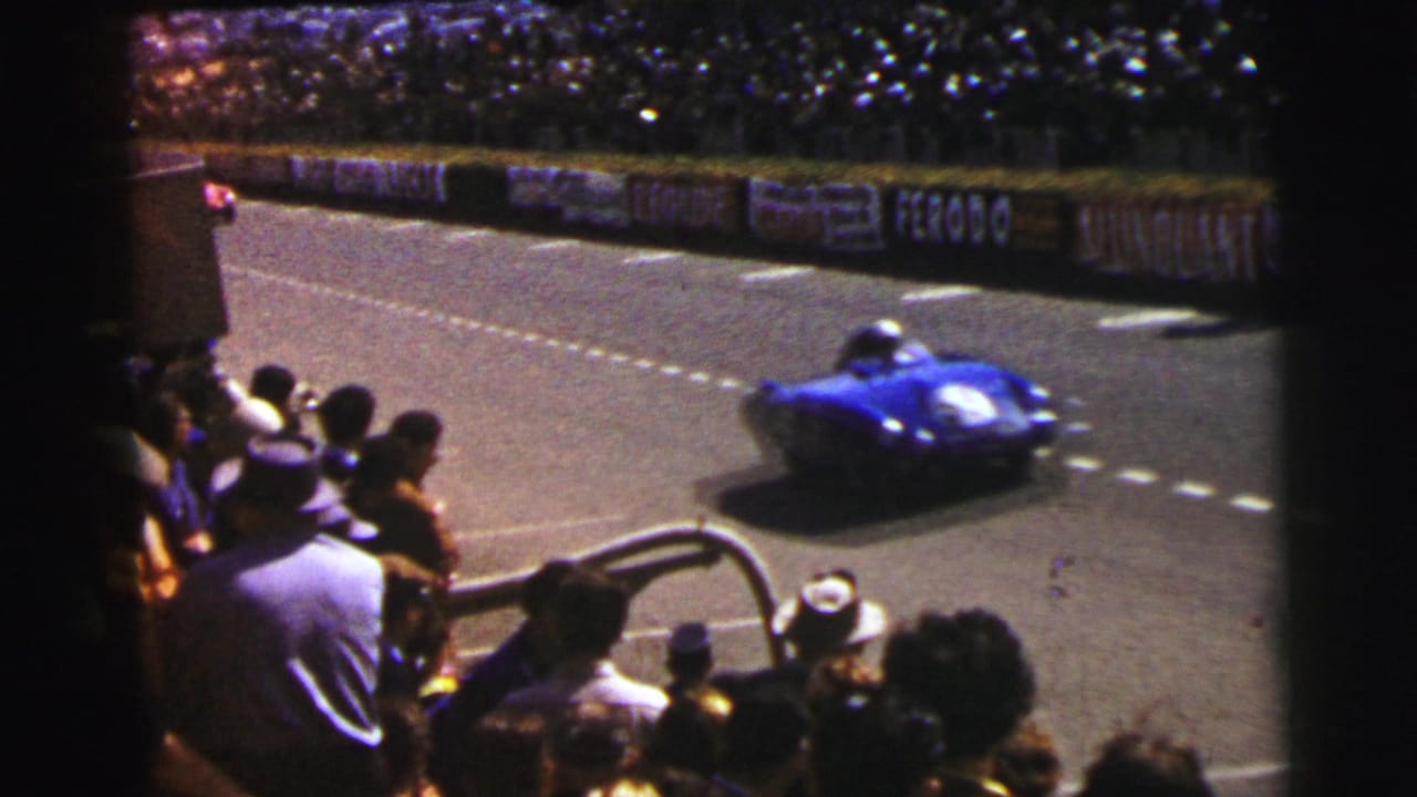 At Le Mans, France, a blue 1950s race car speeds past spectators watching from behind a barrier.