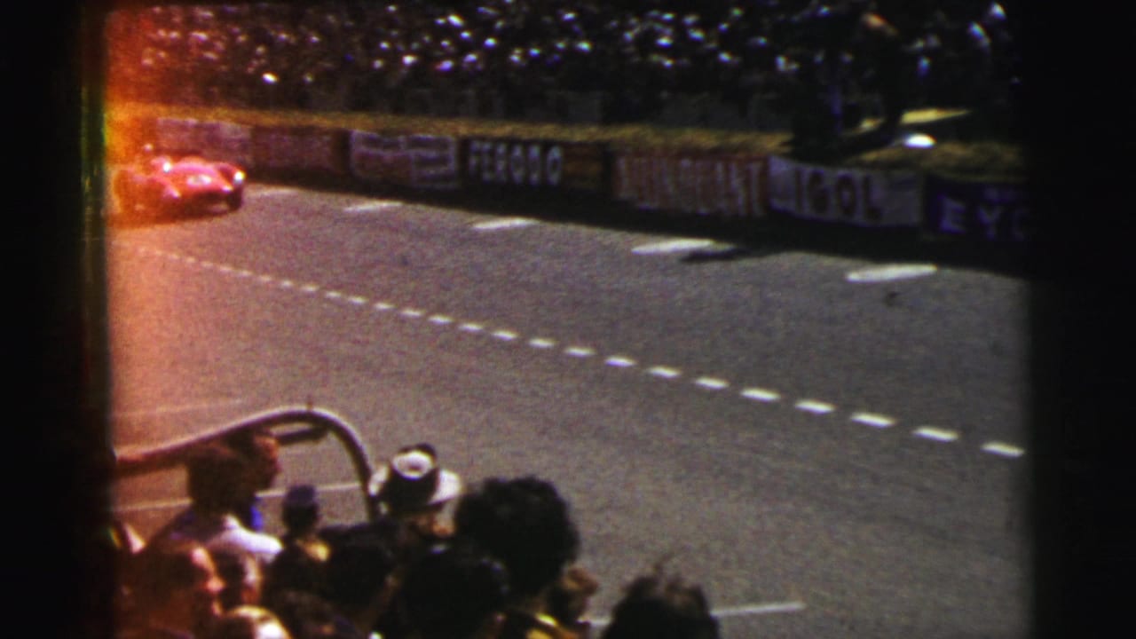 Red race car approaches along the track near sponsor boards and spectators.