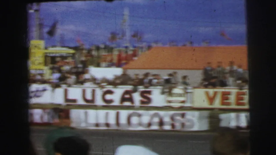 A soft 8mm view of Le Mans trackside stands shows spectators behind large painted advertising boards.