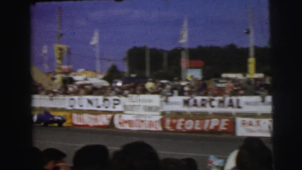 A blue race car passes at the lower edge of the Le Mans frame beneath grandstand signage and spectators.