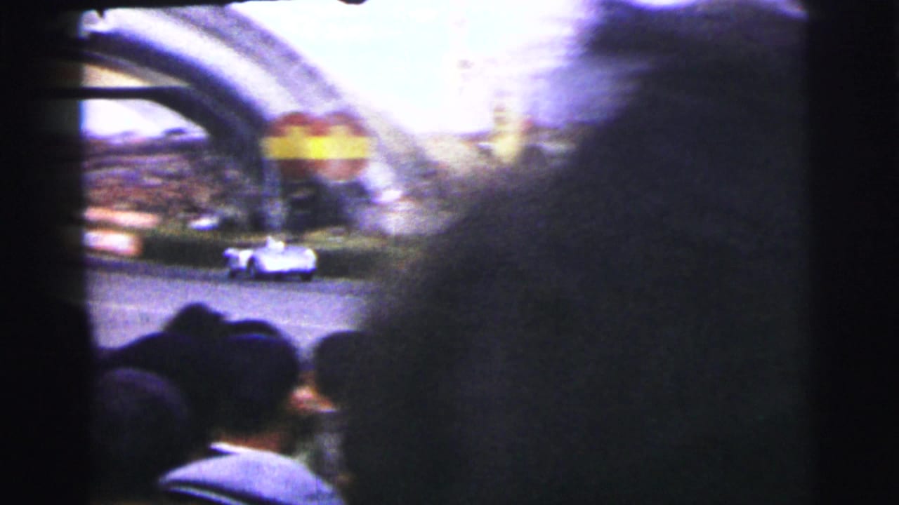 A white race car passes the Dunlop arch area at Le Mans, partly obscured by nearby spectators.