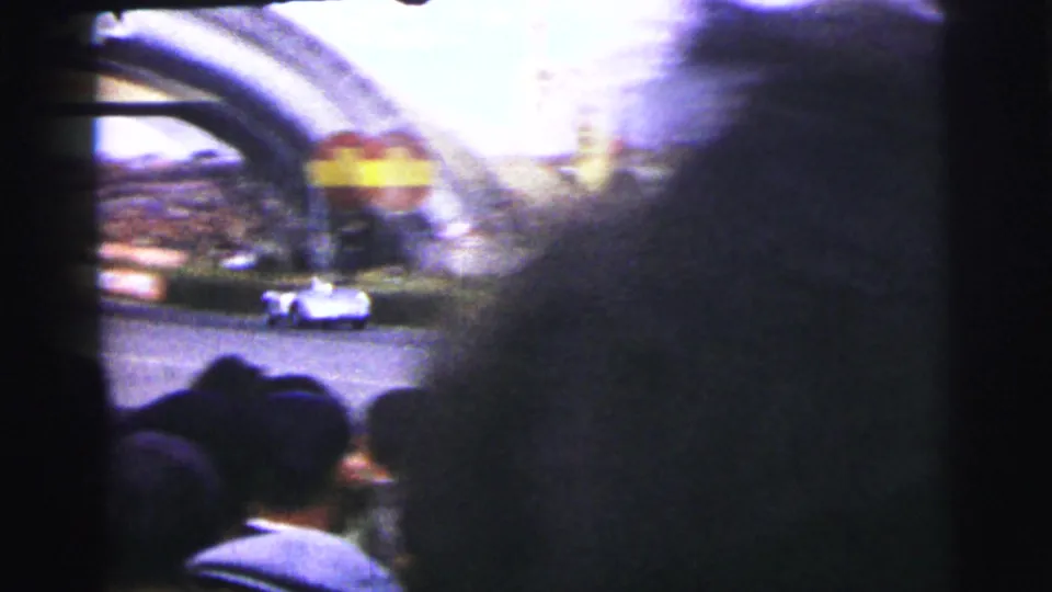 A white race car passes the Dunlop arch area at Le Mans, partly obscured by nearby spectators.