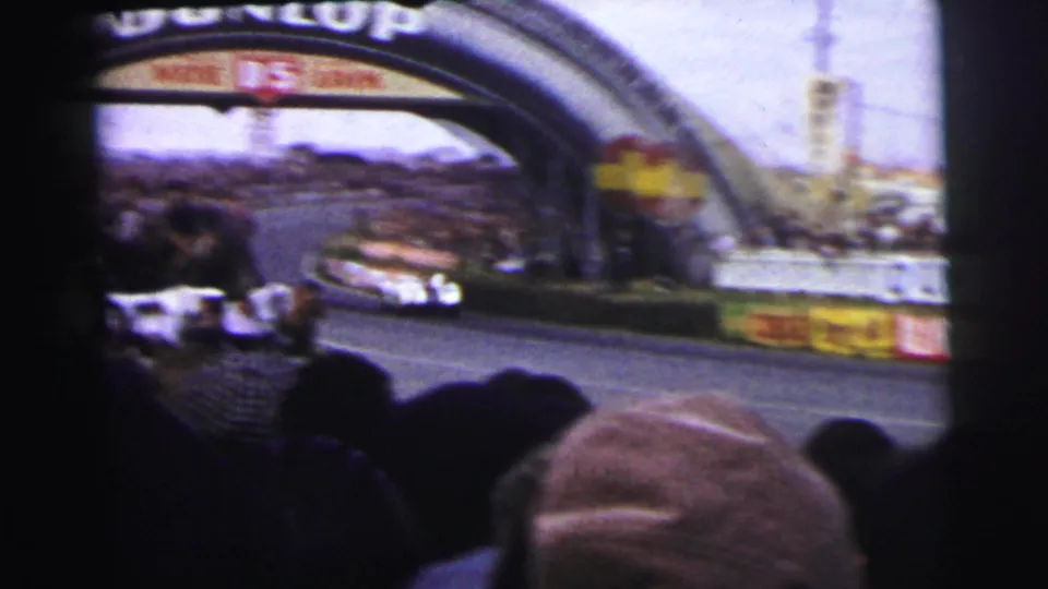 Classic cars pass through the Dunlop bridge section at Le Mans, filmed through heads in the spectator area.
