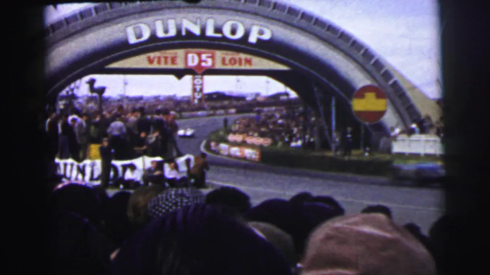 Trackside spectators at 1954 Le Mans watch cars approach beneath the large Dunlop bridge sign.