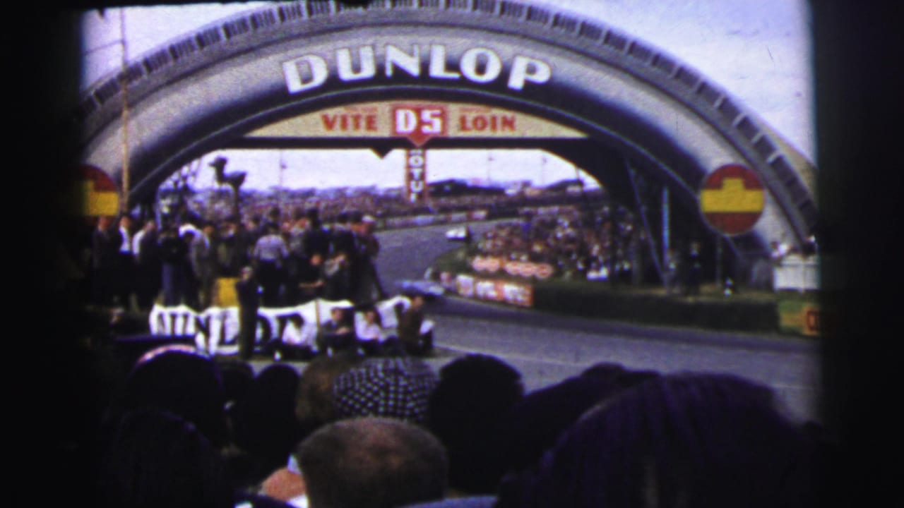 1954 8mm view from behind spectators as cars race beneath the Dunlop arch at Le Mans, France.