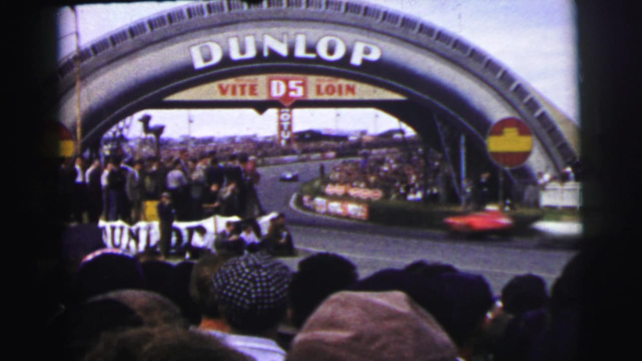 A red race car blurs past spectators under the Dunlop arch in 1954 Le Mans home-movie footage.