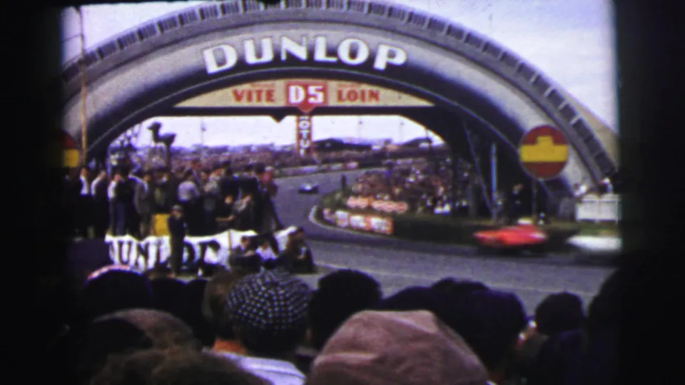 A red race car blurs past spectators under the Dunlop arch in 1954 Le Mans home-movie footage.