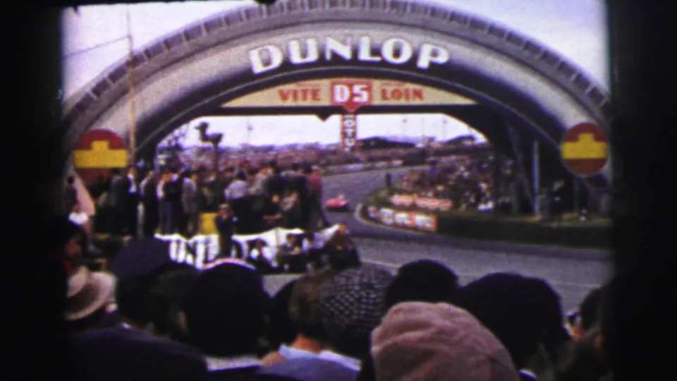 Crowded roadside view of a dark racing car moving beneath the Dunlop arch at Le Mans in 1954.