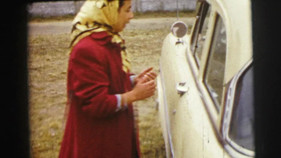A woman in a red coat stands next to a light-colored car, likely part of the same 1954 home-movie reel.
