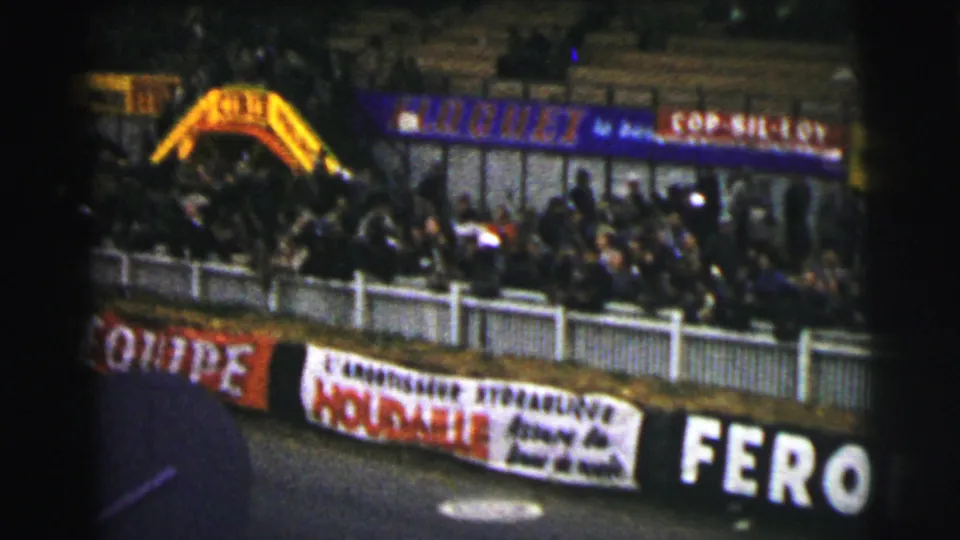 1954 Le Mans crowd along the barrier, with Pirelli-style trackside advertising and a yellow arch in the background.