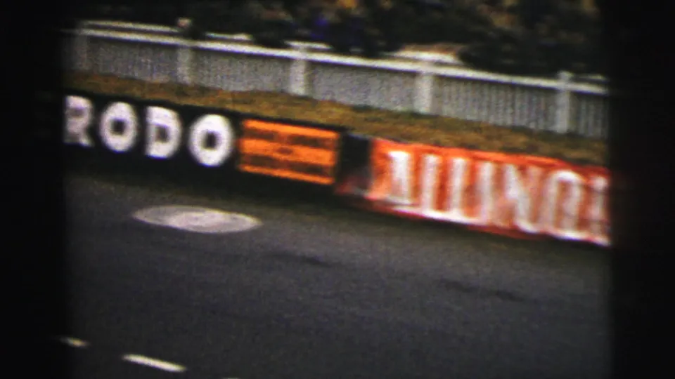 1954 Le Mans trackside view with roadway, white barrier, and blurred commercial boards along the raceway edge.