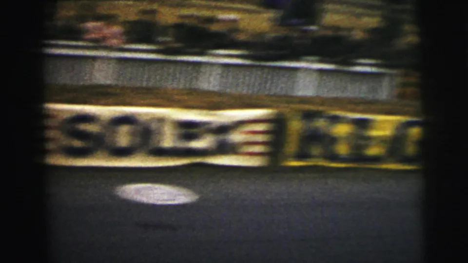 Trackside signage at Le Mans appears behind a safety barrier, with the view blurred by fast camera movement on 8mm film.