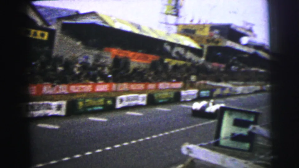 Le Mans race track view shows grandstands, spectators behind barriers, advertising boards, and a small race car on the circuit.