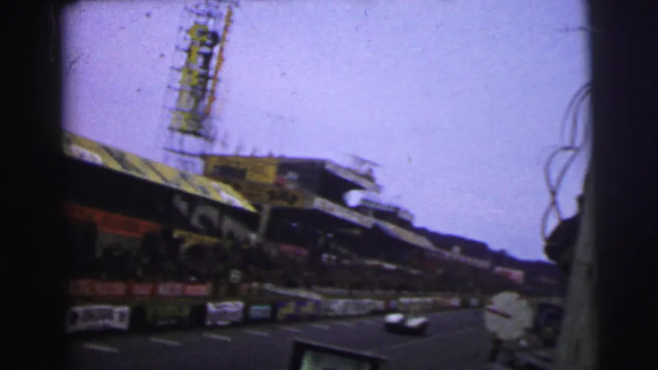 Wide 1954 Le Mans track view with a grandstand, tower structure, advertising boards, barriers, and a race car moving through the frame.