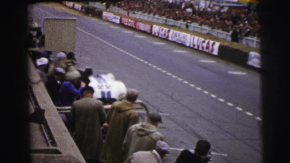 A white race car with dark striping appears beside the pit-side spectators in this 1954 Le Mans home-movie frame.