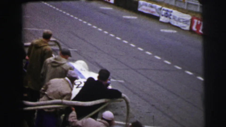 The white number 25 race car stops or slows near the Le Mans pit rail with spectators close by in period clothing.