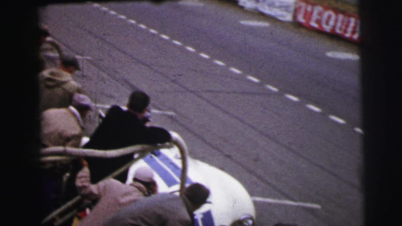 At Le Mans in 1954, men gather near a white and blue open racing car stopped beside dashed track markings.