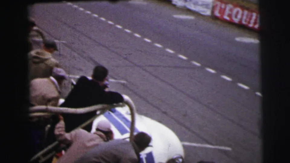 At Le Mans in 1954, men gather near a white and blue open racing car stopped beside dashed track markings.