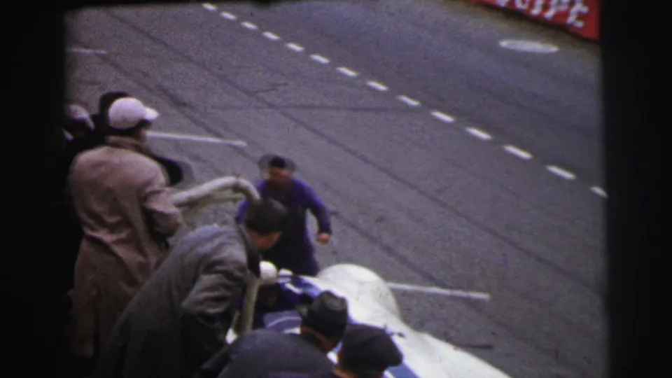 At the 1954 Le Mans circuit, several men cluster around the cockpit of a white and blue racing car on the track.