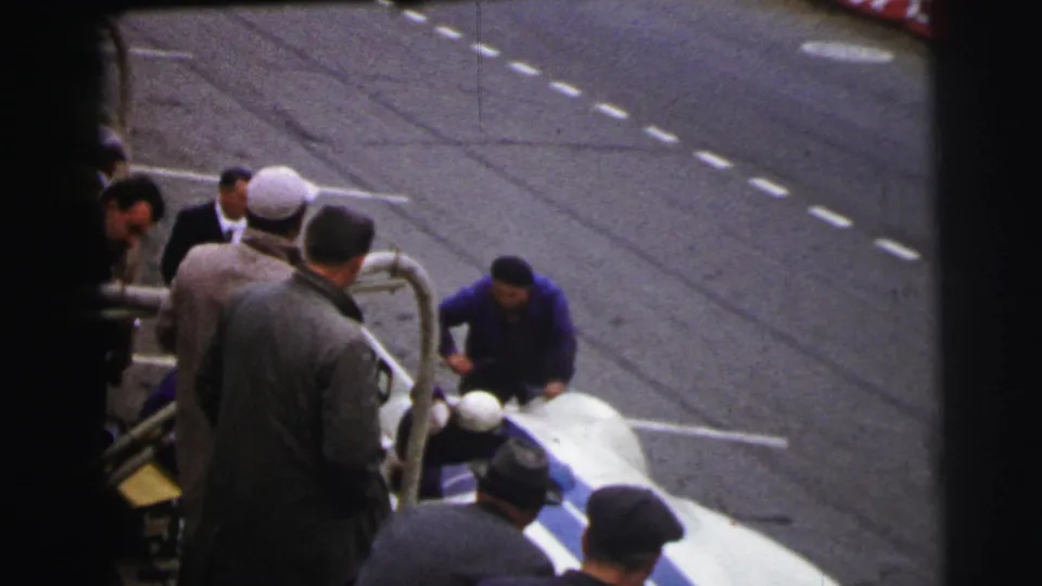 A man bends toward the stopped white and blue car as other adults watch from a close trackside position at Le Mans in 1954.