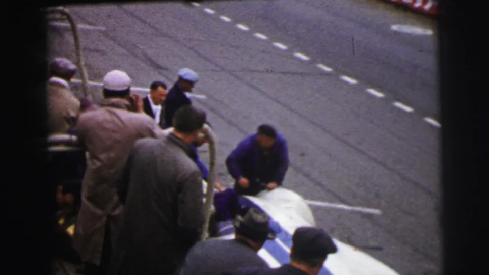 Men in coats and caps stand close to a stopped racing car on the Le Mans circuit, with red banners visible beyond the asphalt.