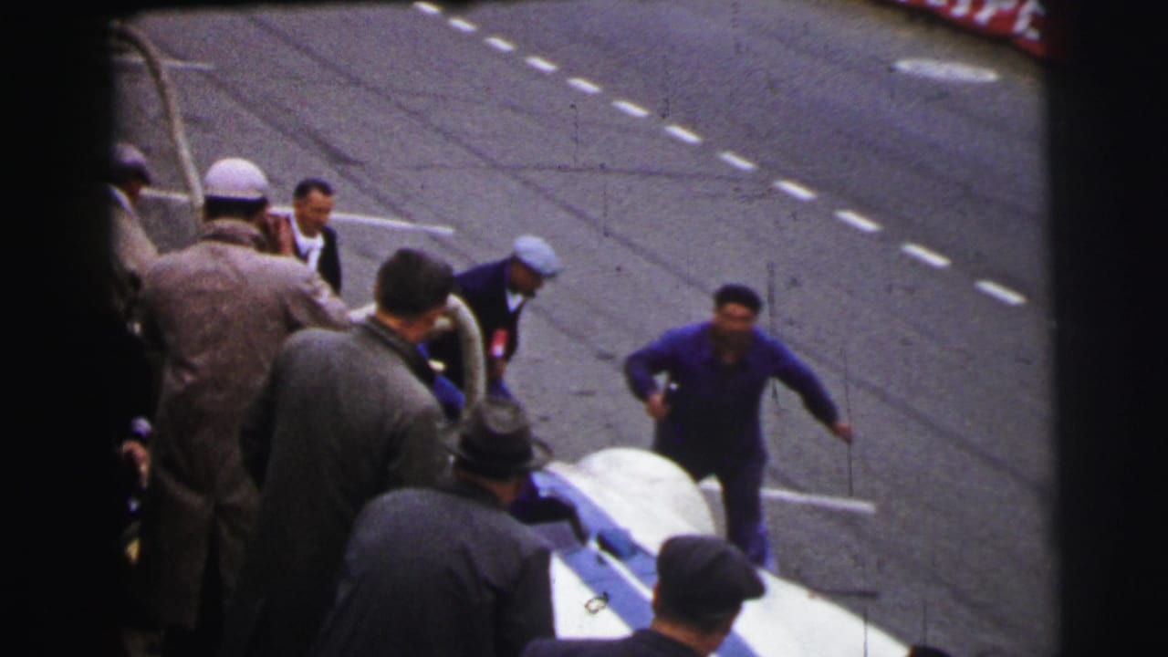 At Le Mans in 1954, mechanics in blue overalls and nearby spectators cluster around a white and blue race car during a pit stop.