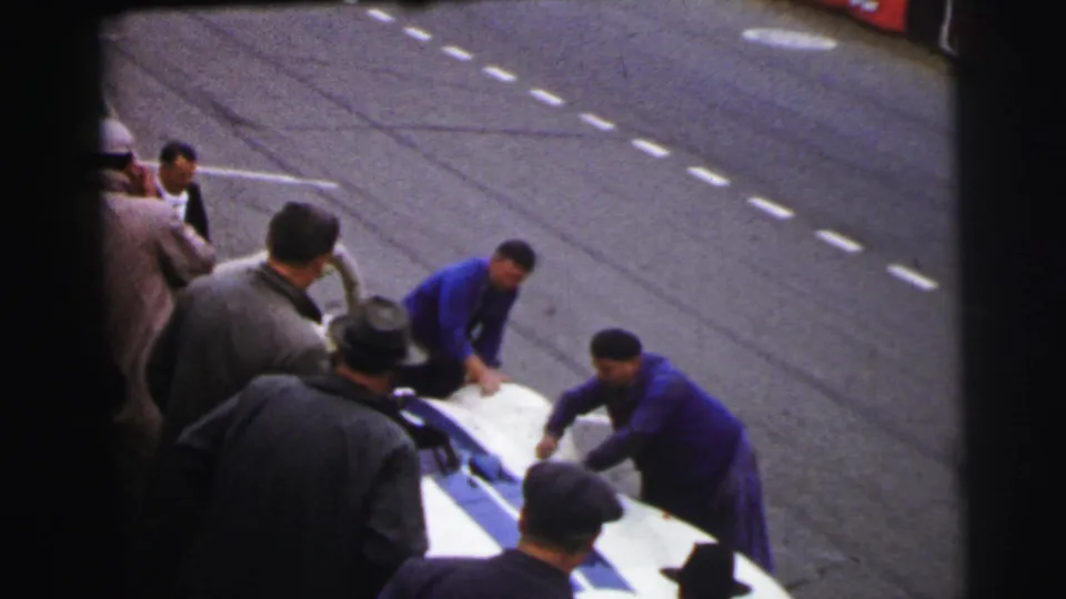 At the 1954 Le Mans setting, two mechanics in blue overalls work at the front of a white and blue race car as observers stand close by.