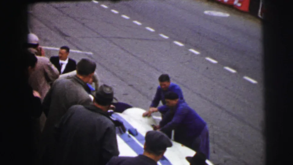 A 1954 Le Mans pit-stop view shows a mechanic bent over the race car with suited men and cap-wearing spectators watching nearby.
