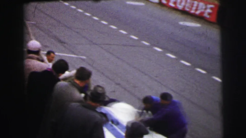 The closing frame shows mechanics still bent over the white and blue car at Le Mans, with spectators at left and marked asphalt behind them.