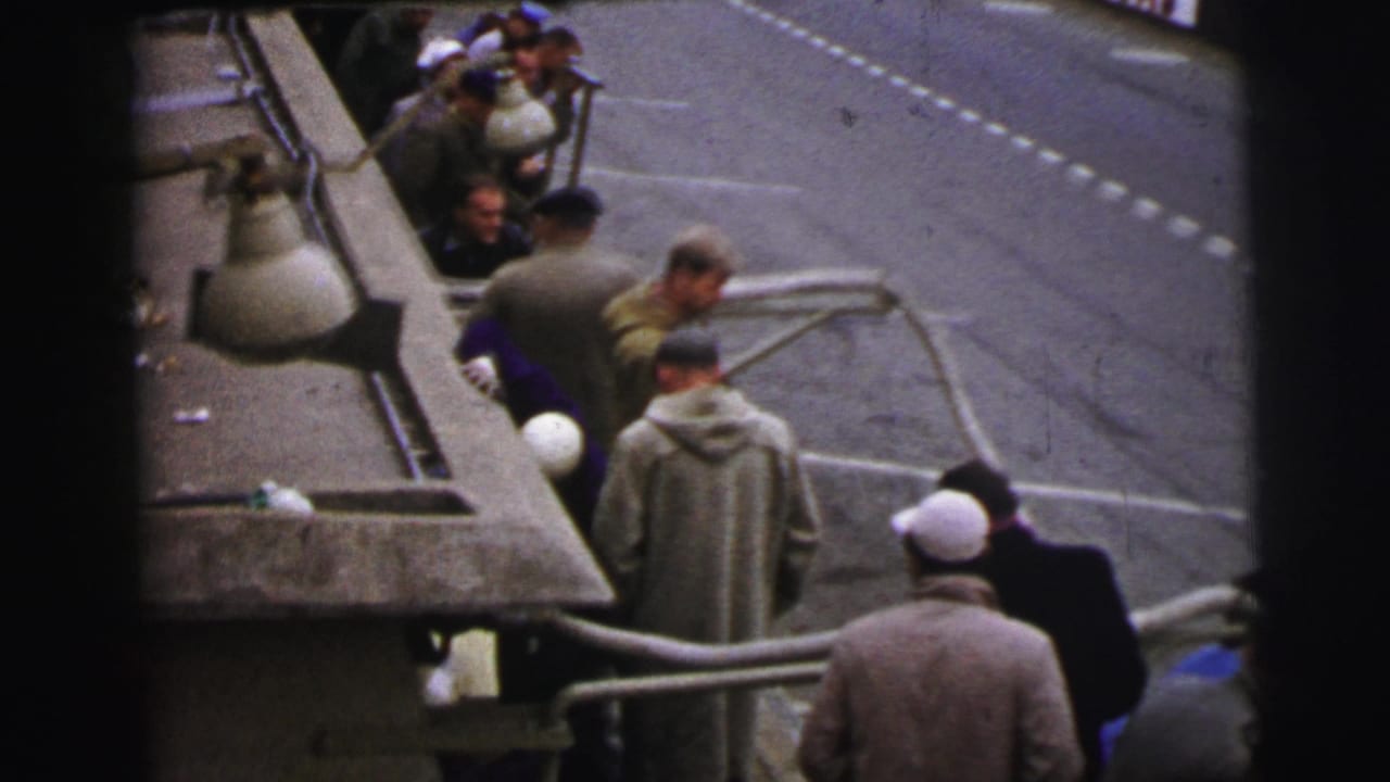 At Le Mans in 1954, men in period coats and caps cluster beside a race car at the pit edge while the track markings are visible nearby.