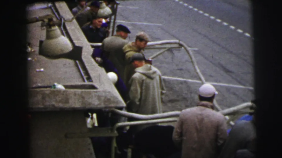 Men in mid-century clothing stand around a race car in the Le Mans pit area, with concrete barriers and painted racing surface behind them.