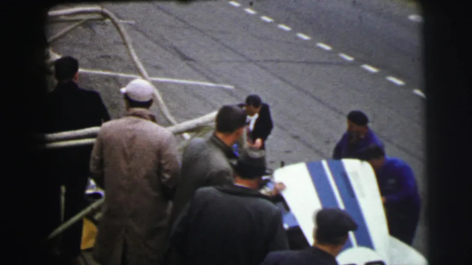 At the 1954 Le Mans pit area, a man appears to lift open bodywork on a race car while several men in coats and caps watch or assist.
