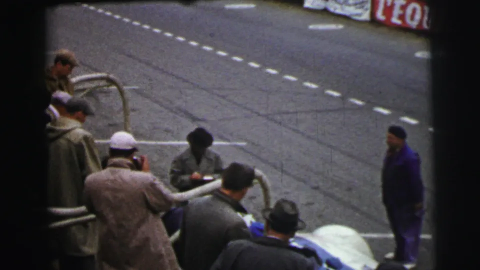 The Le Mans pit edge remains busy as men in coats and caps stand near the car and a man in blue walks along the track side.