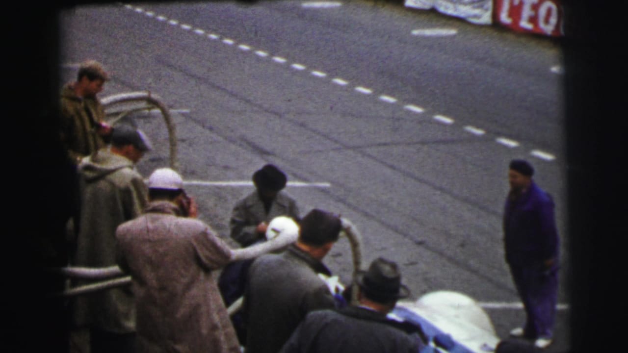 Men in coats and hats gather by metal railings beside a race track.