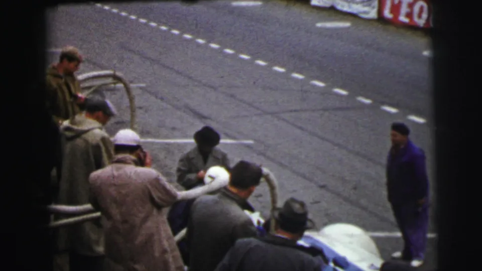 At Le Mans in 1954, bundled spectators stand near metal barriers beside the asphalt track, with papers and a pale race car partly visible.