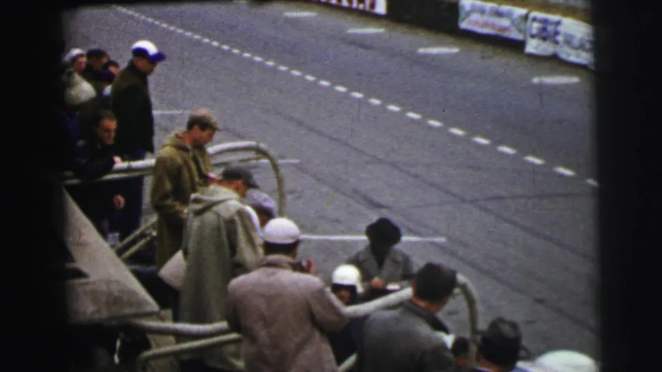 A 1954 Le Mans trackside view shows men in heavy coats and hats gathered at railings, with papers handled near a partially seen car.