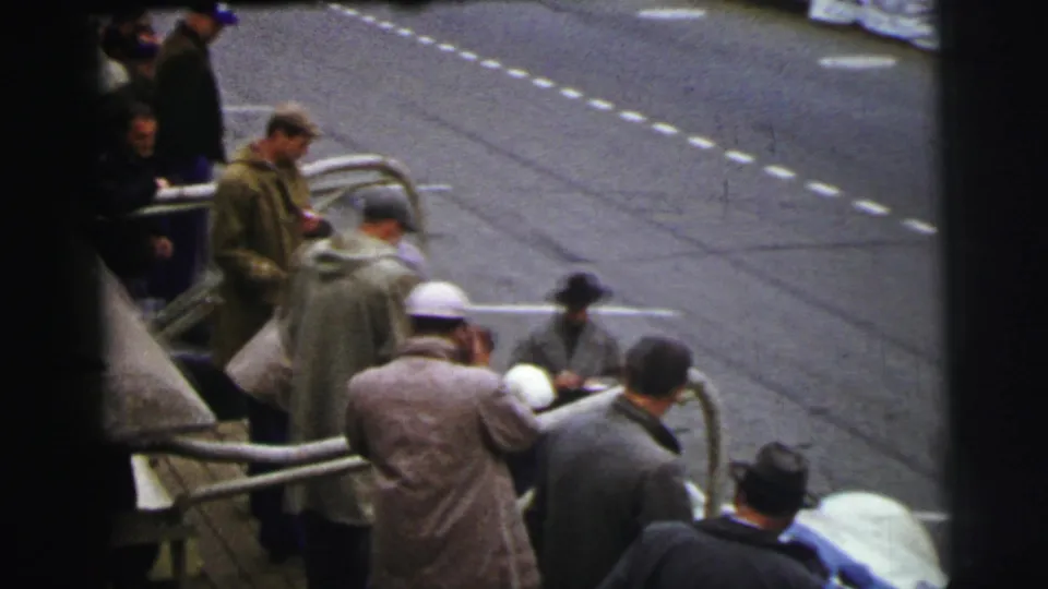 The 1954 Le Mans footage shows bundled men at a metal barrier, seen from above with dashed track lines and a partial race car in the foreground.