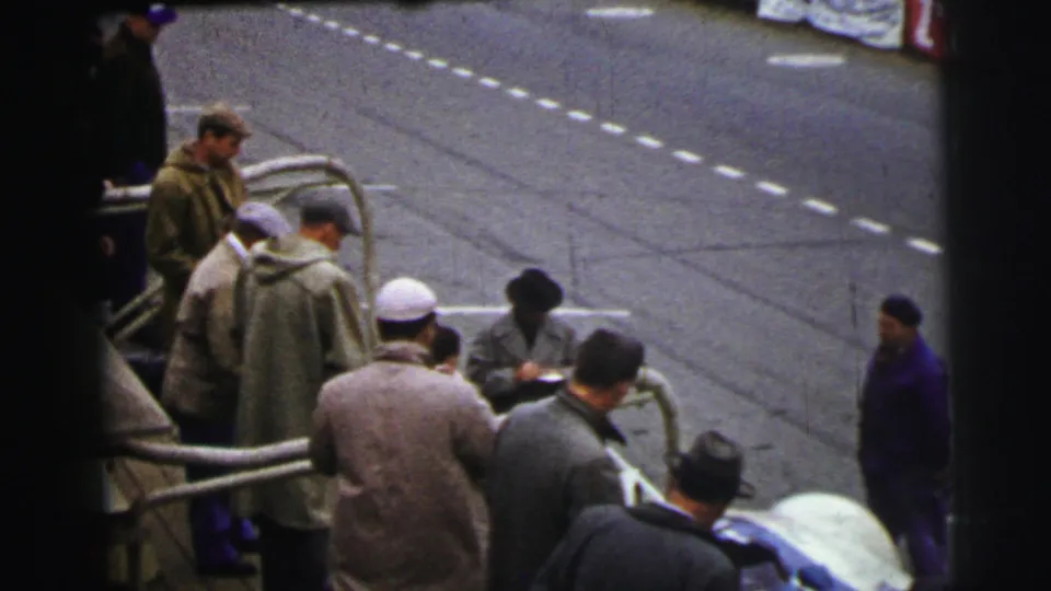 At the Le Mans race setting in 1954, spectators in coats and hats remain gathered at the rail as the empty marked track stretches behind them.