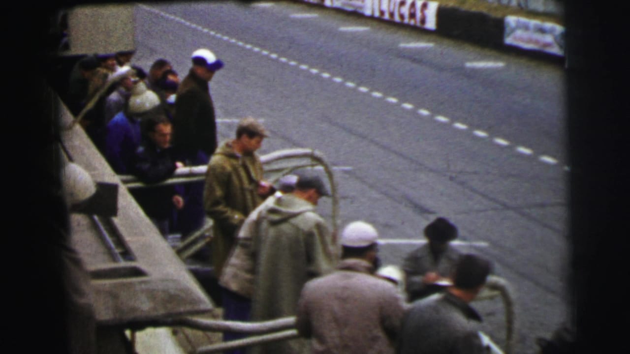 Spectators in coats and hats stand behind a roadside barrier beside a race track.