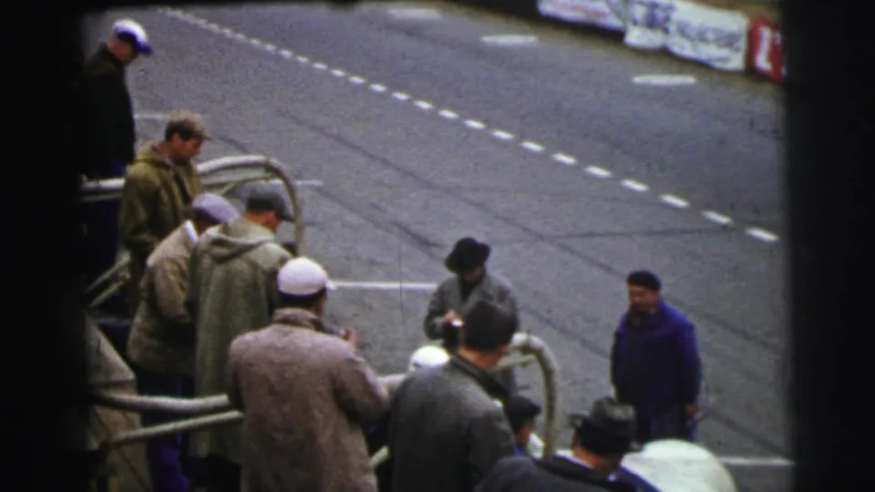 Men at the Le Mans trackside barrier look toward the road, with dashed markings and advertising banners visible beyond them.
