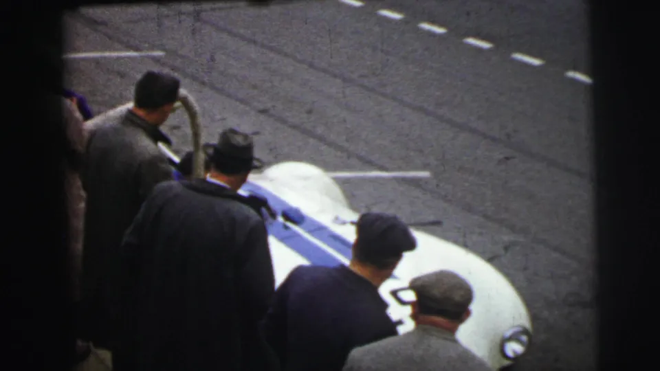A white race car with blue striping appears near the Le Mans trackside crowd, partially framed by spectators in coats and hats.