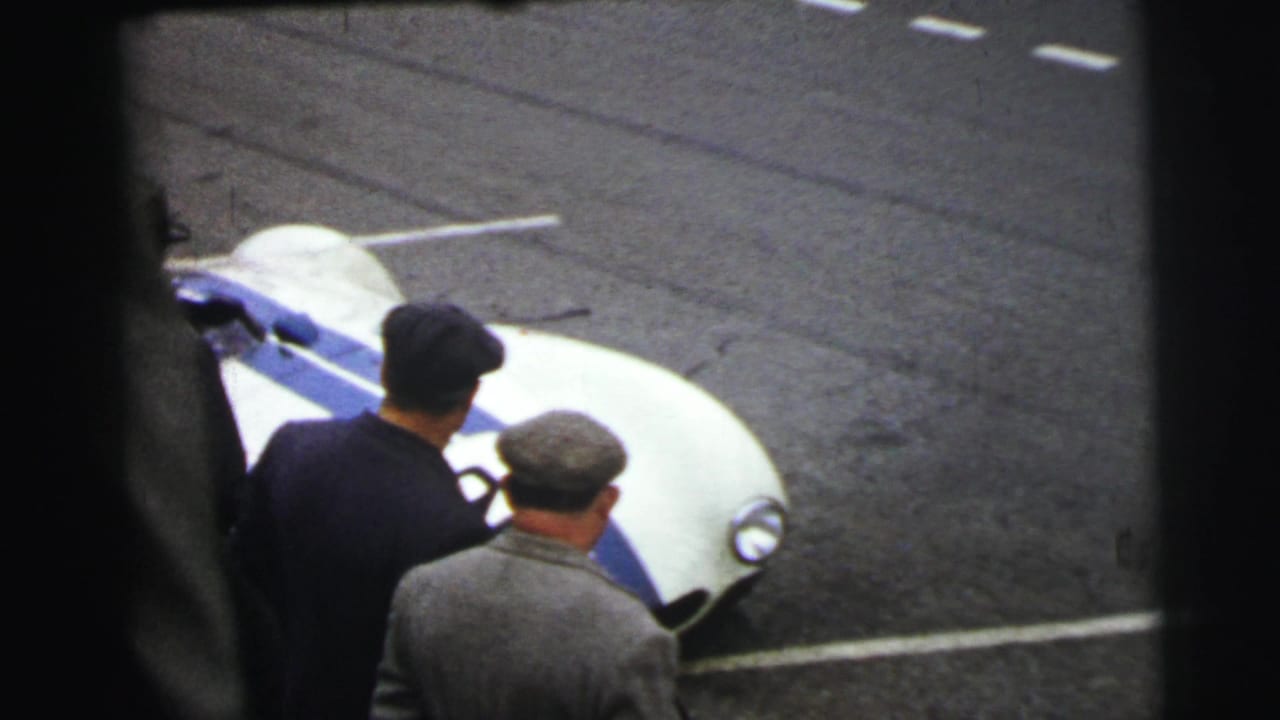 At Le Mans in 1954, men in coats and hats stand beside the front of a white race car with blue striping and number 92.