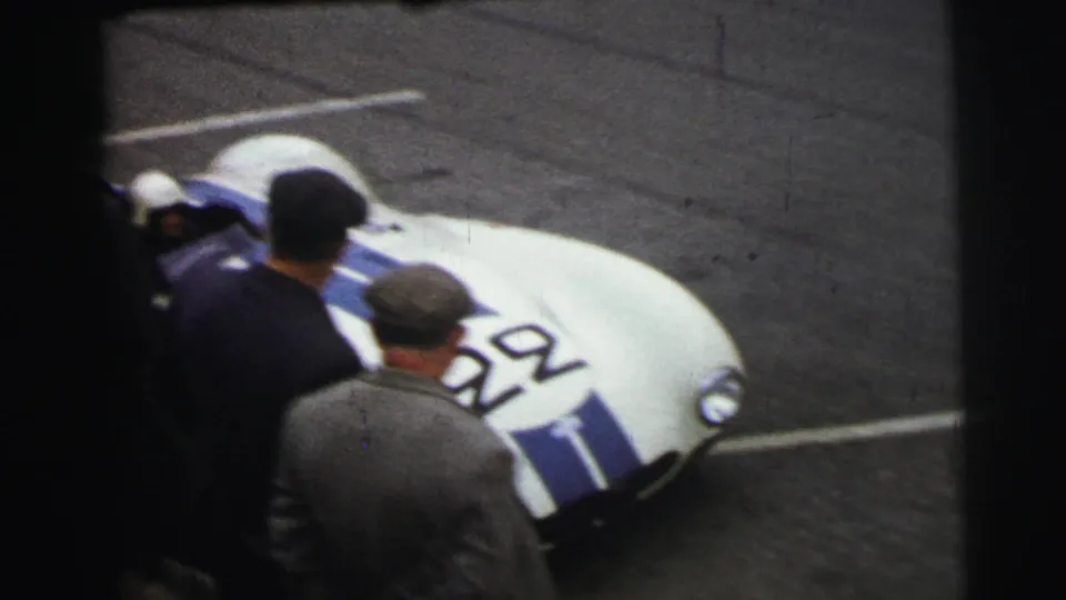 At Le Mans in 1954, a white car with blue stripes and number 92 is seen from above with men standing close by.