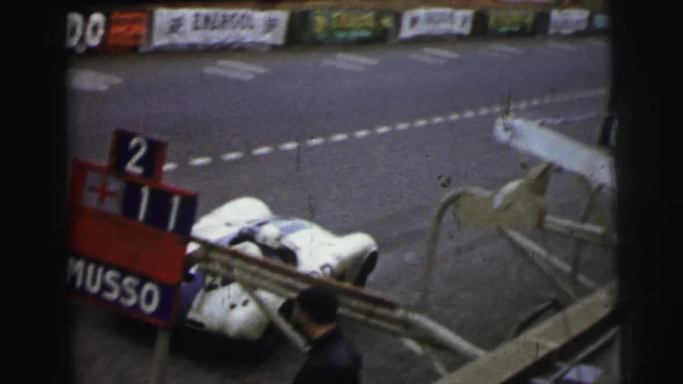 White race car by trackside barriers at Le Mans, with pit board numbers and sponsor signage visible in the background.