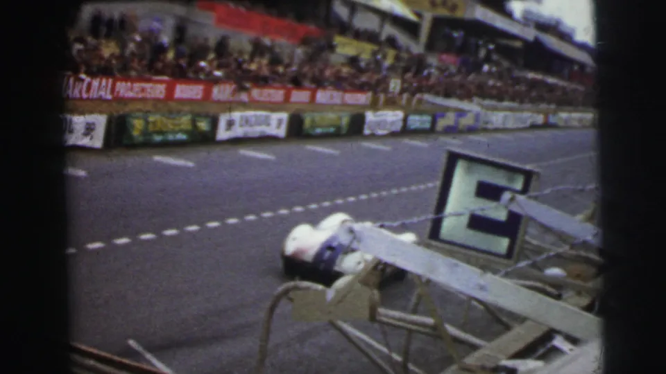 Le Mans race car viewed from beside the circuit, with crowded stands, barriers, signs, and a large pit board in frame.
