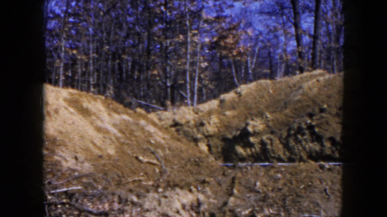 1954 Hicksville home-movie frame showing disturbed earth, a dirt mound, tire ruts, and bare trees beyond the excavation area.