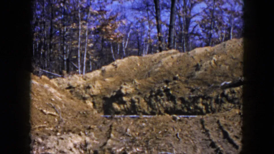 1954 Hicksville frame with an exposed dirt bank, tire-marked ground, bare trees, and a possible pipe or rod across the soil.