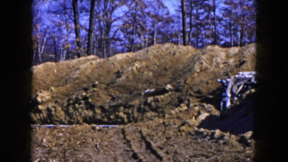 1954 Hicksville frame showing disturbed soil, tire ruts, trees, and a small partial wooden structure or debris near the right edge.