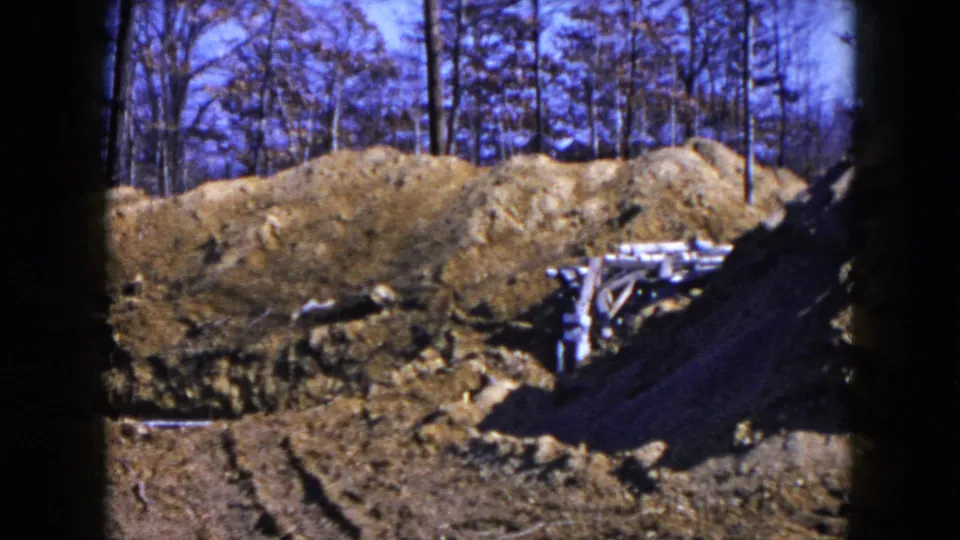 1954 Hicksville construction-site frame with raised earth, loose boards, and a wooded backdrop under blue sky.