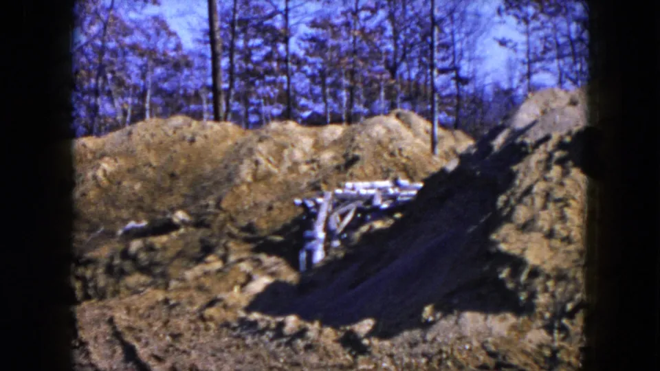 8mm home-movie frame from the Hicksville site showing an excavated lot, wood debris, and bare trees beyond the dirt piles.